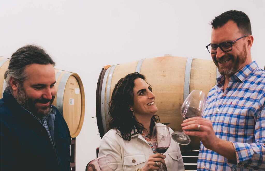 Allison, Kieran, and Josh smiling at a wine tasting with wine barrels in the background
